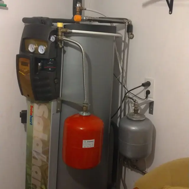 Depósito solar vertical dentro de casa, com central de comando ao lado, um vaso de expansão vermelho e outro cinzento.
