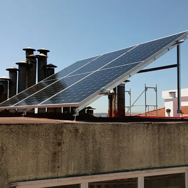Painel fotovoltaico montado em estrutura inclinada num terraço, ao lado de um conjunto de chaminés.